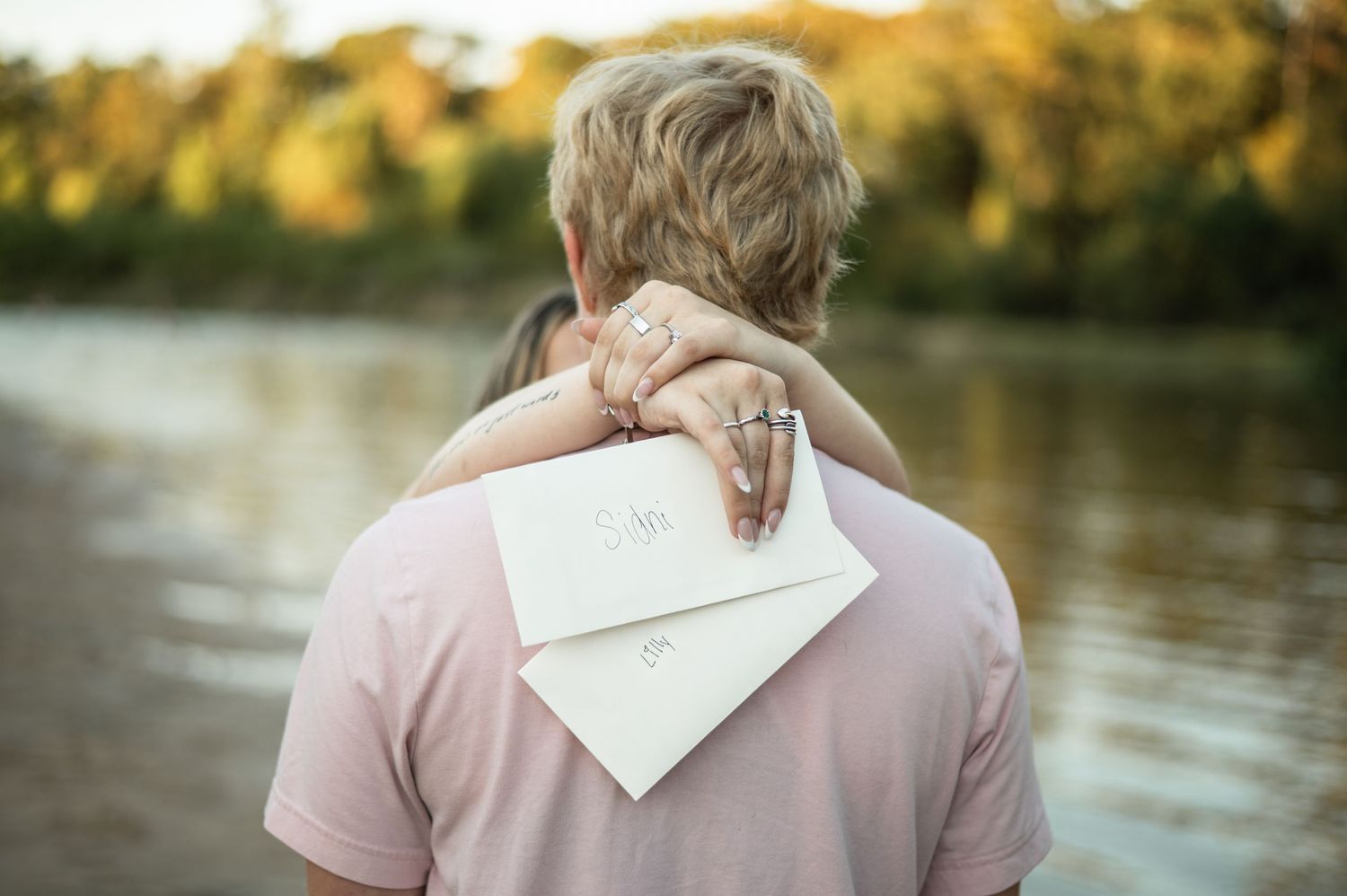 Couple in soft pink and white outfits embrace by a tranquil lake during sunset.