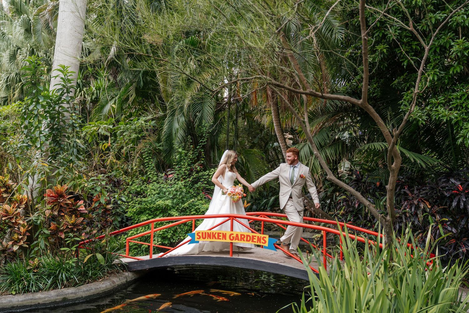 A couple walks across a charming wooden bridge surrounded by lush tropical gardens and palm trees.