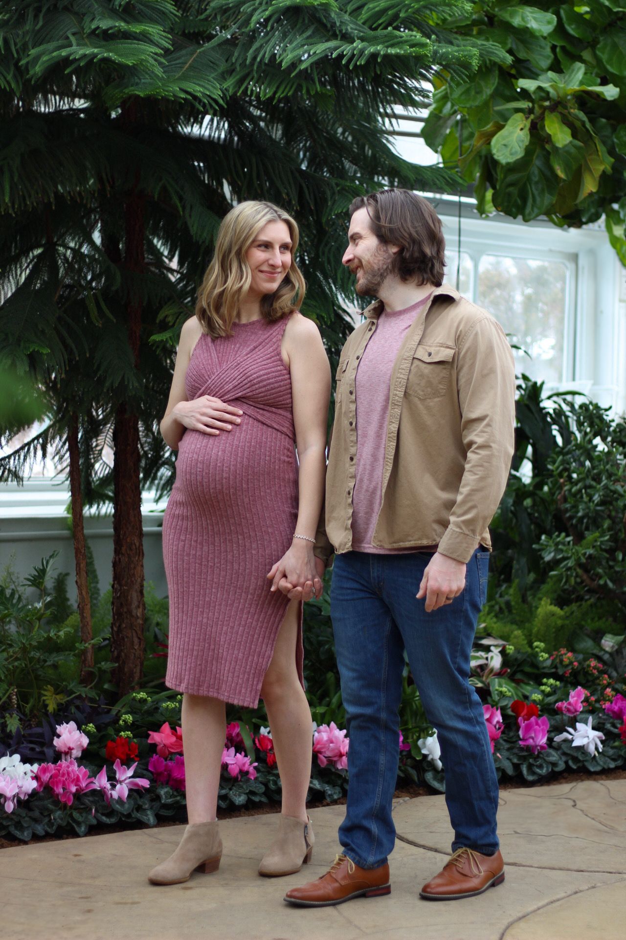 Couple poses together in greenhouse with pink flowers and palm trees in background.