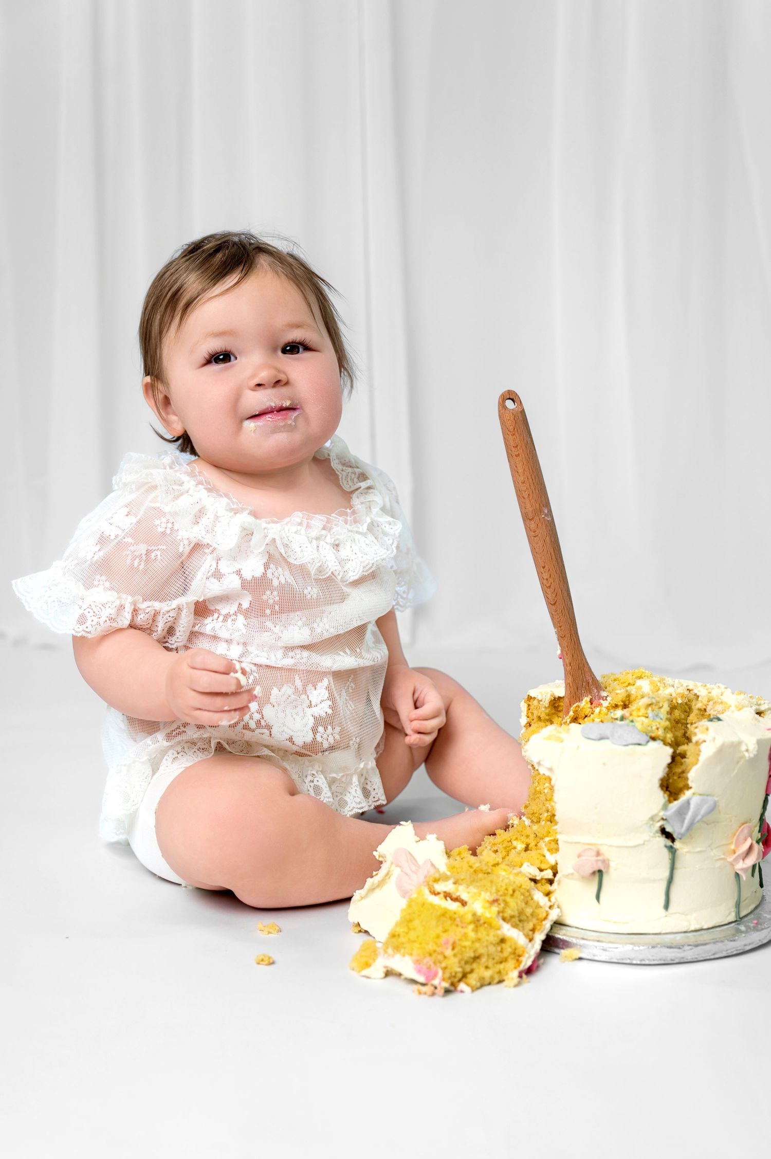 A small child in white lace outfit sits next to a smashed birthday cake with gold decorations on white background.