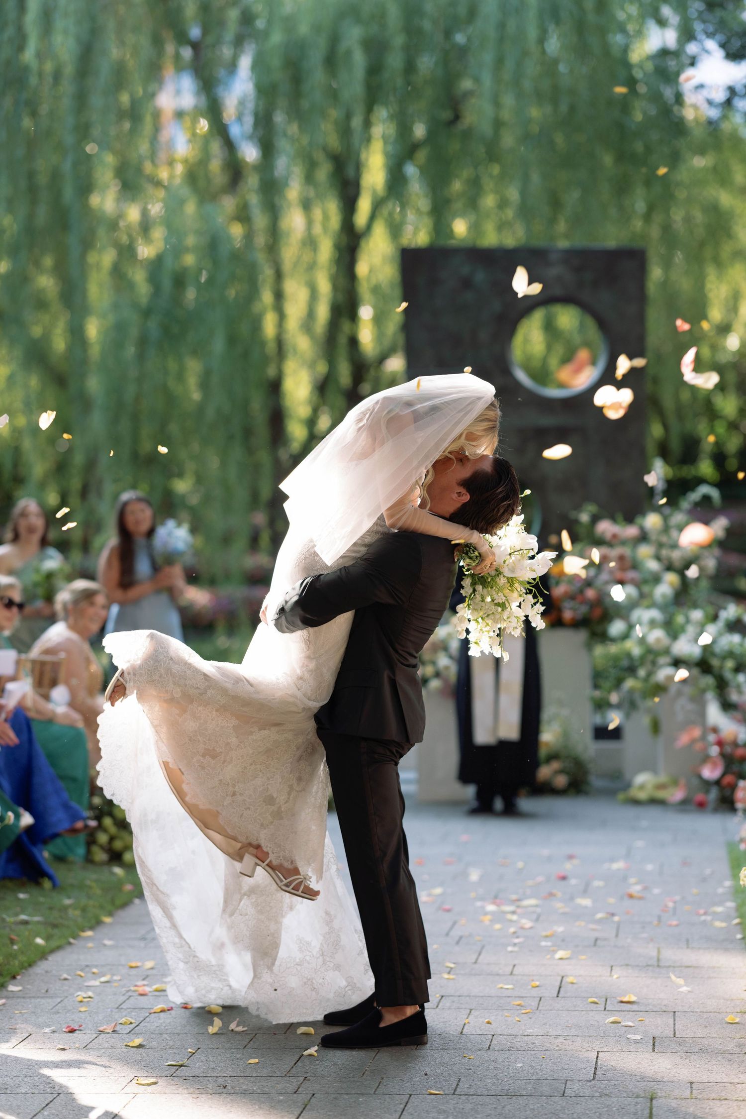 A wedding couple dances romantically beneath weeping willow trees in a garden setting with falling flower petals.