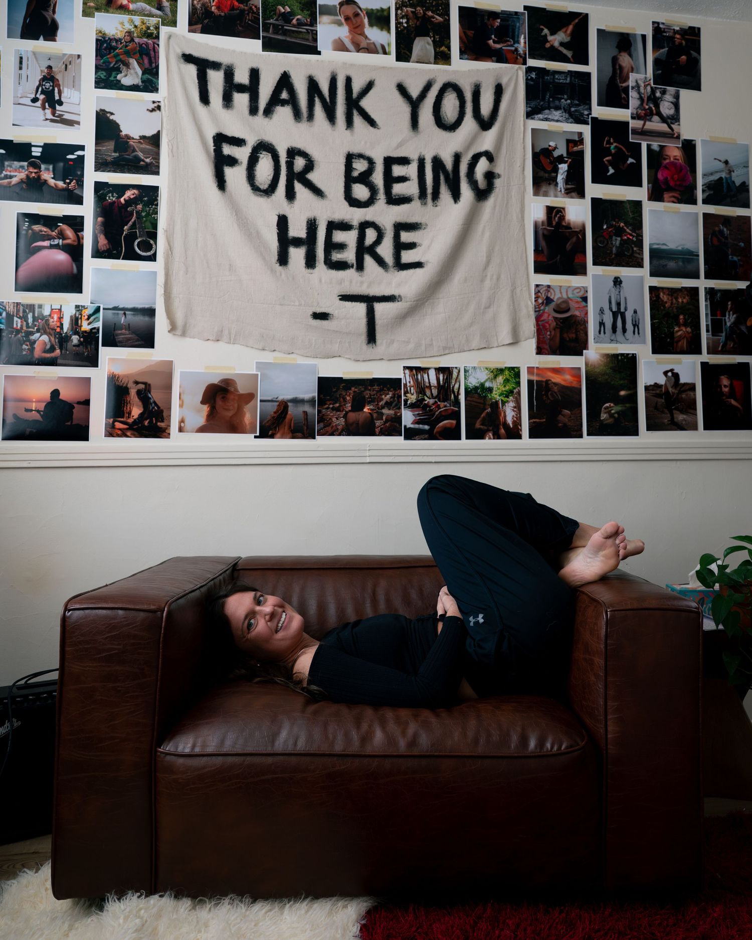 A brown leather couch sits beneath a wall covered in photographs and a banner with text reading 'THANK YOU FOR BEING HERE'.