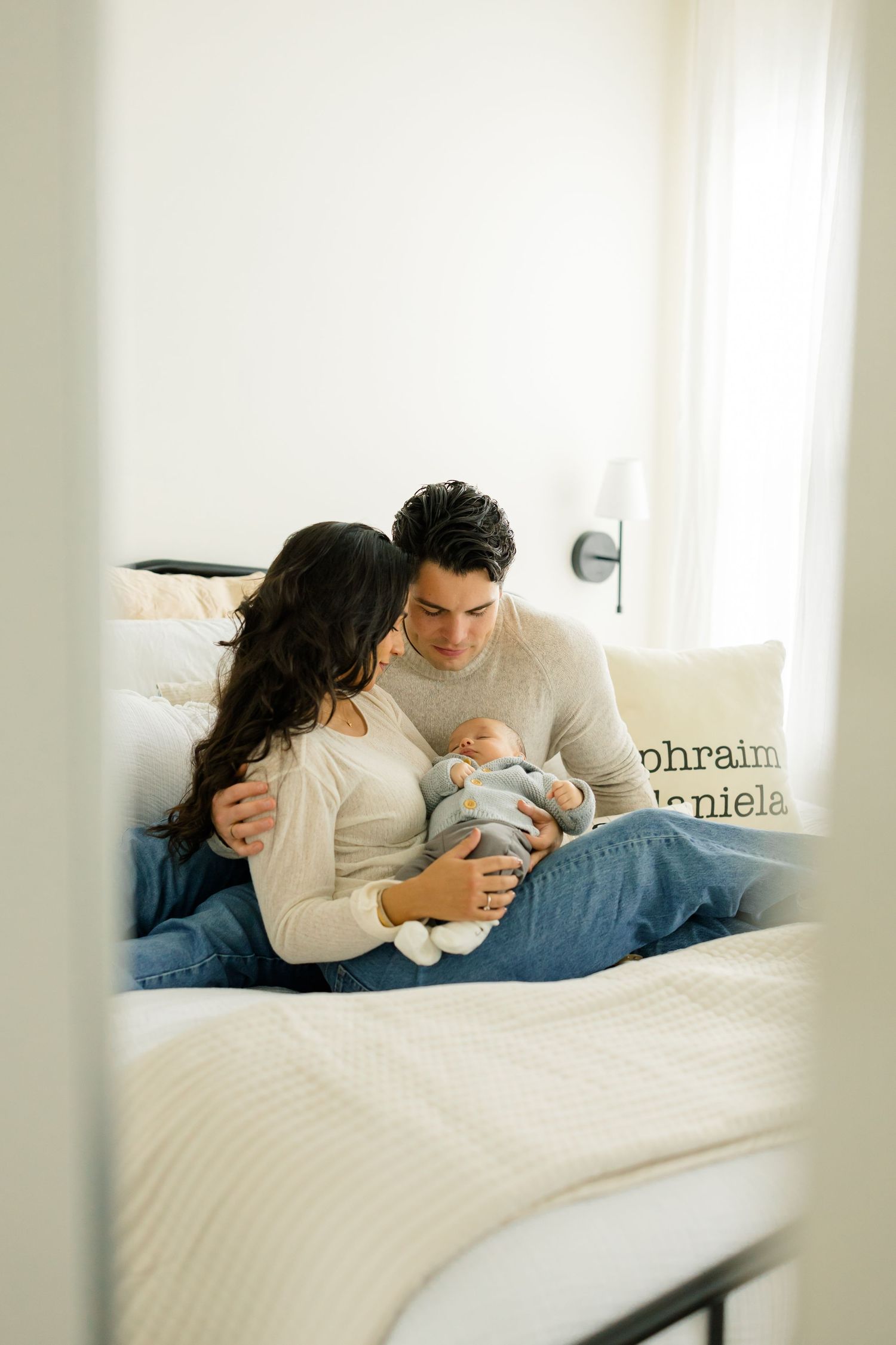 A couple relaxes together on a white bed while looking at their phones in natural light.