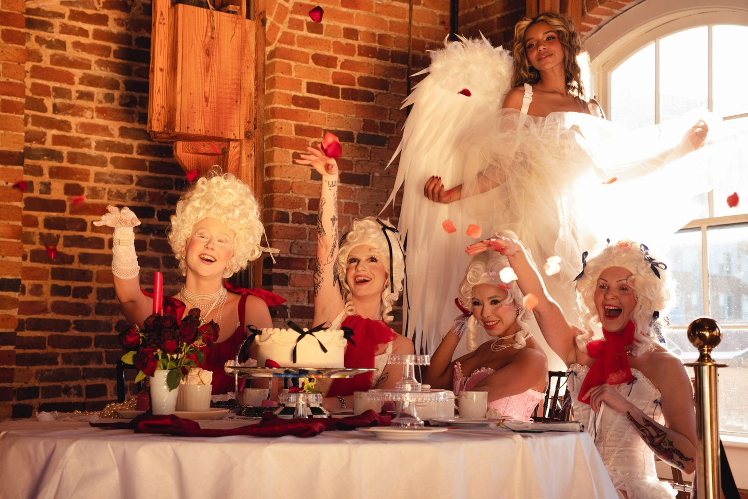 A festive tea party scene with elegant white wigs and feathers at a rustic brick venue with vintage decor.