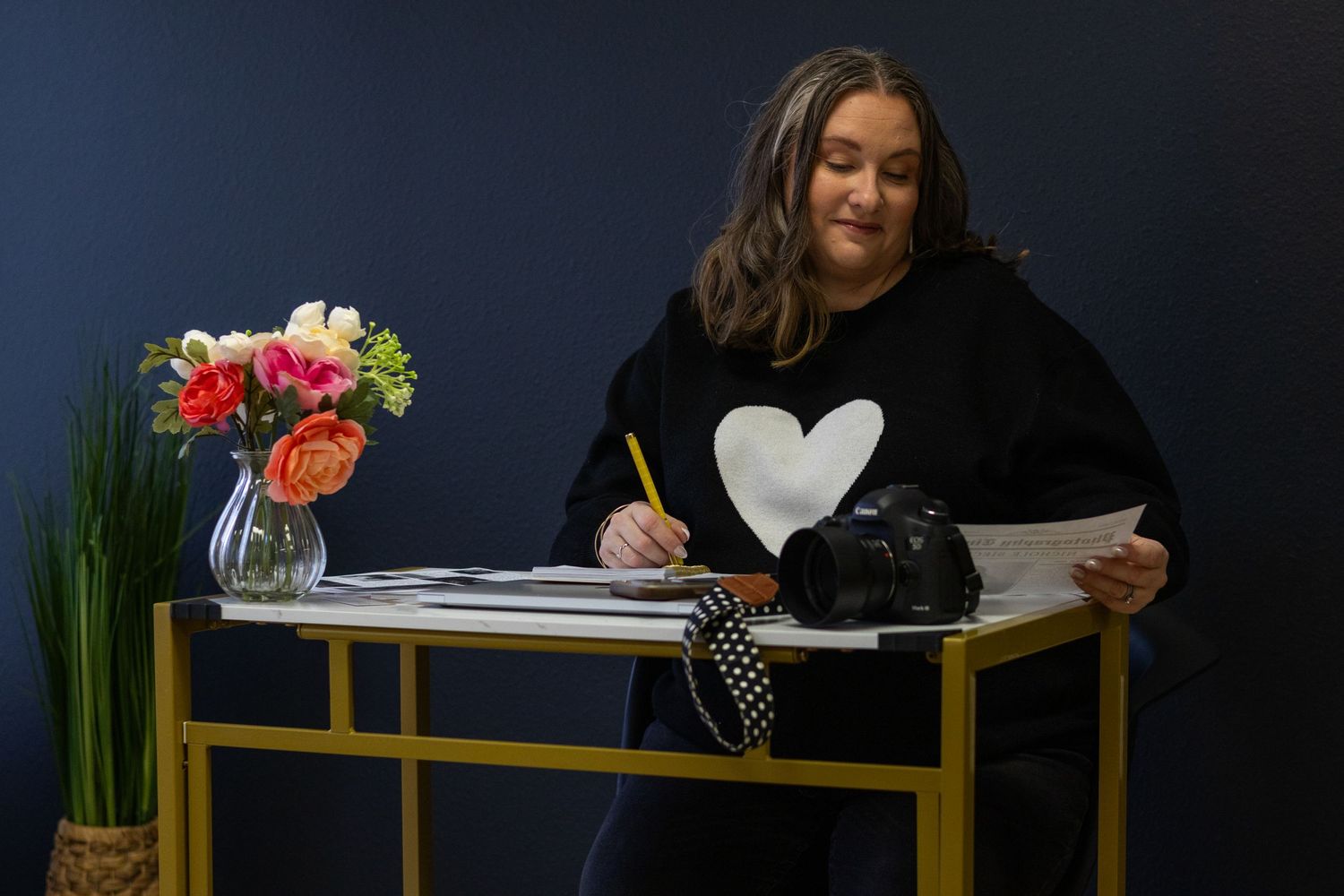 Someone wearing a black sweater with heart design writes at a stylish gold desk with floral arrangement.
