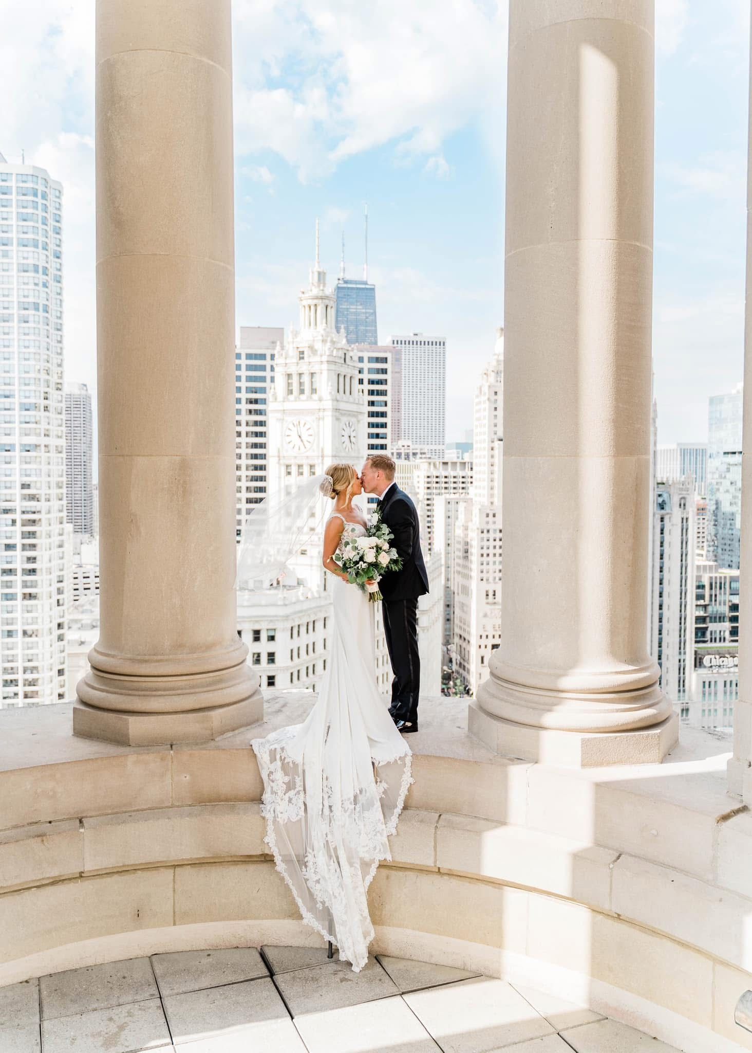 A romantic wedding couple embrace between elegant white columns overlooking a city skyline on a bright sunny day.