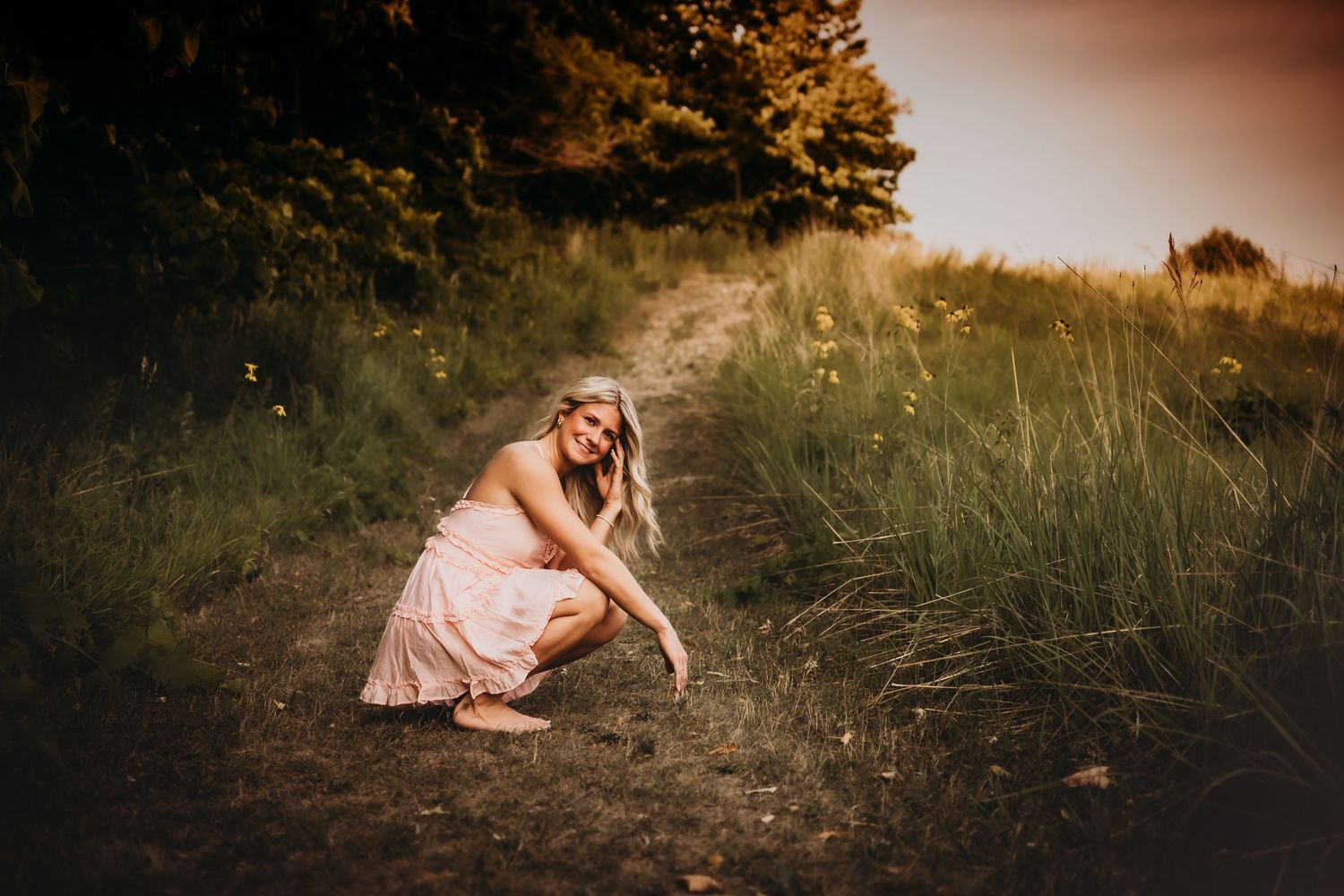 Person in pink dress crouches on a dirt path surrounded by tall grass at golden hour.