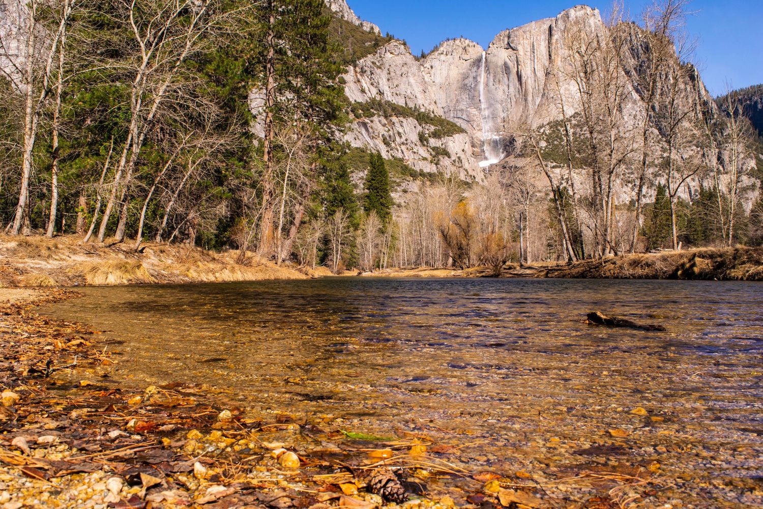 Crystal clear river waters flow past autumn leaves with towering granite cliffs in the background.