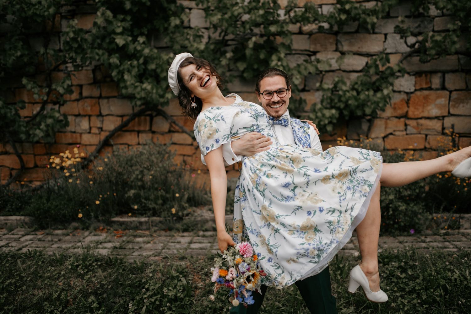 Joyful groom lifting laughing bride in floral dress outside rustic stone wall.