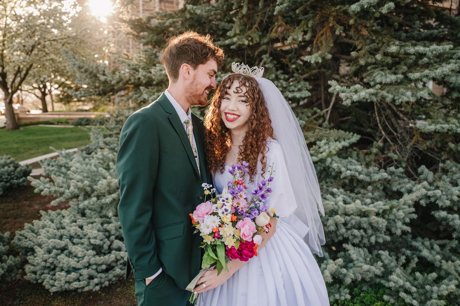 A couple in wedding attire sharing a joyful moment in front of evergreen trees with colorful flowers and a sunlit background.
