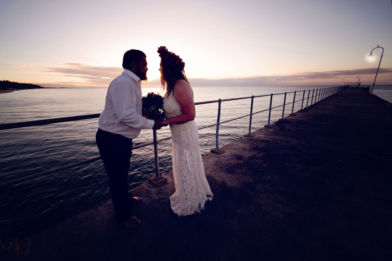 A couple shares a romantic moment on a pier at sunset while standing near the railing over tranquil waters.