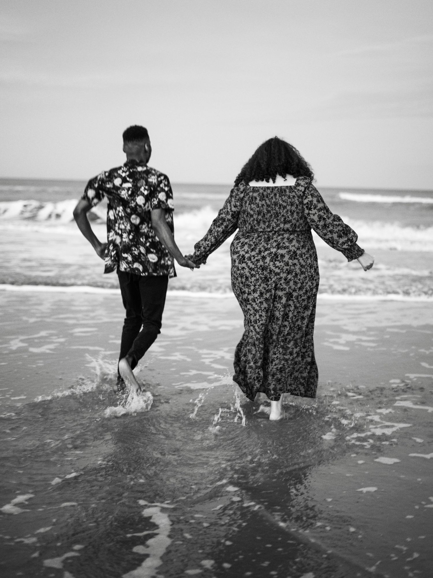 A couple walks hand in hand along the beach shore in patterned clothing, captured in black and white.