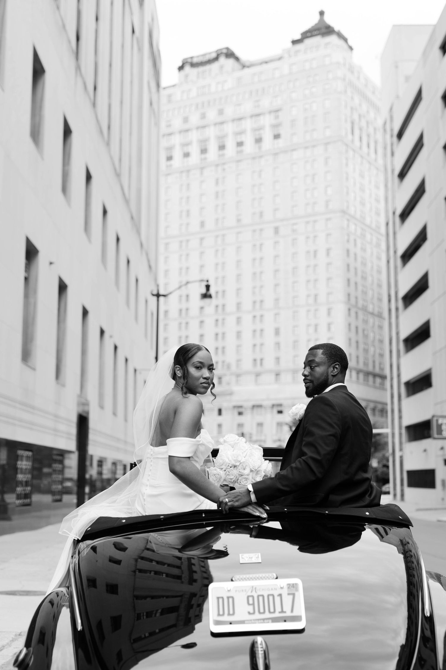 Black and white wedding photo of newlyweds sitting on a vintage car with city buildings in background.
