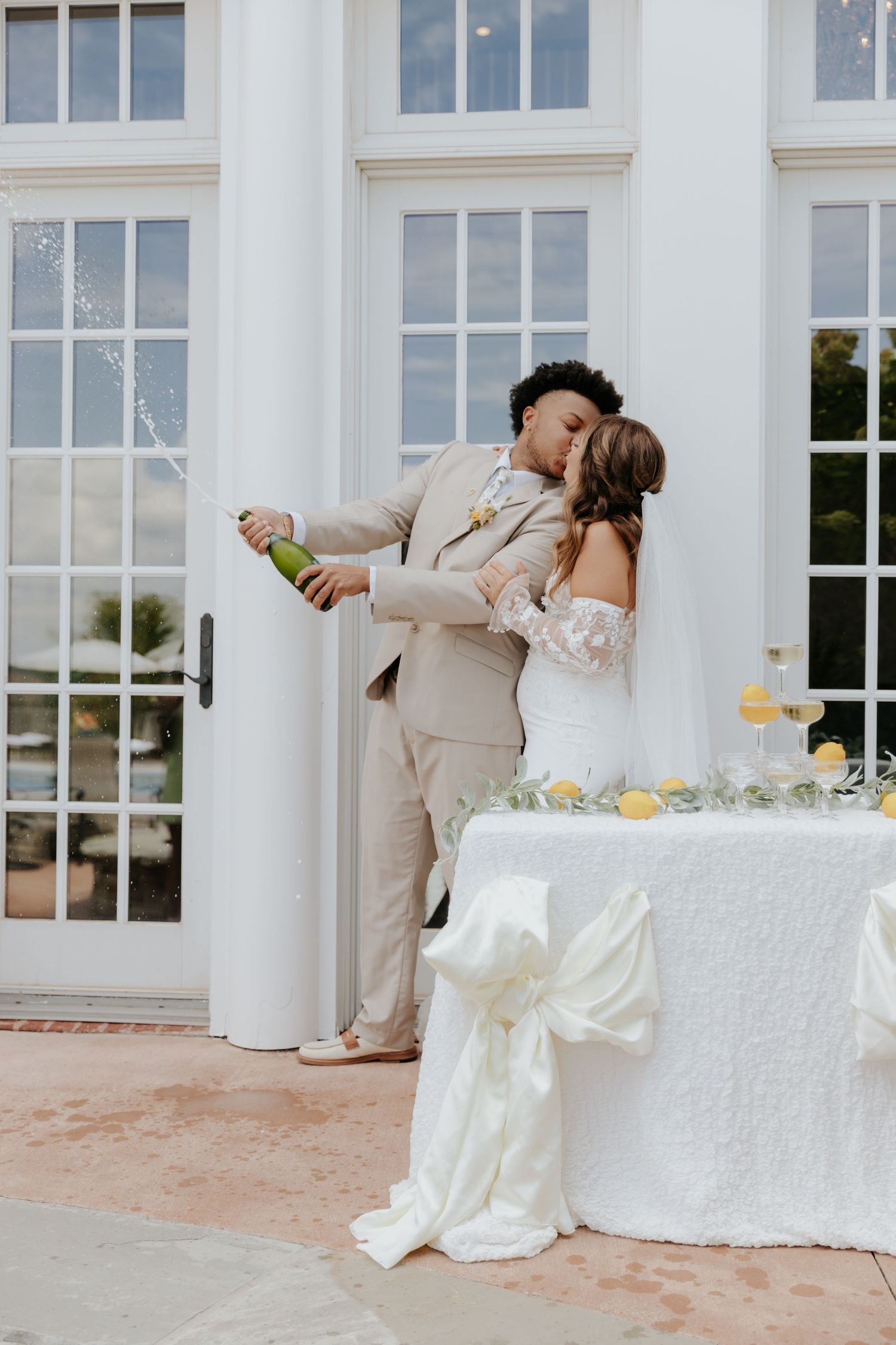 A couple shares a tender moment next to an elegant white wedding table with champagne flutes at a formal venue.