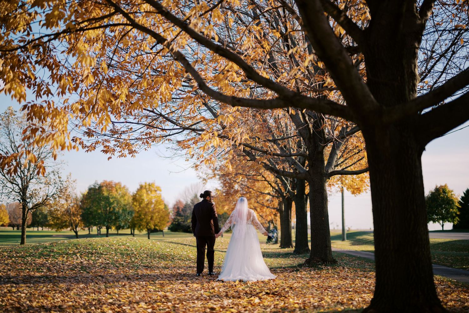 A couple walks beneath golden autumn trees on a leaf-covered path in a serene park setting.