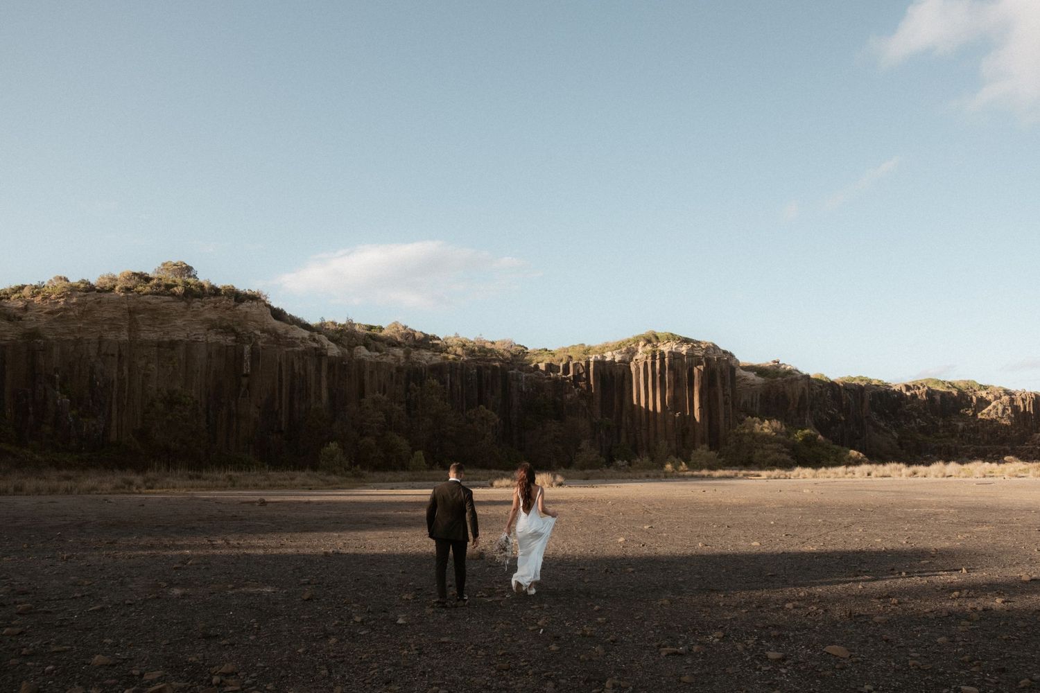 Couple walking hand in hand across desert landscape with cliffs in background at dusk.