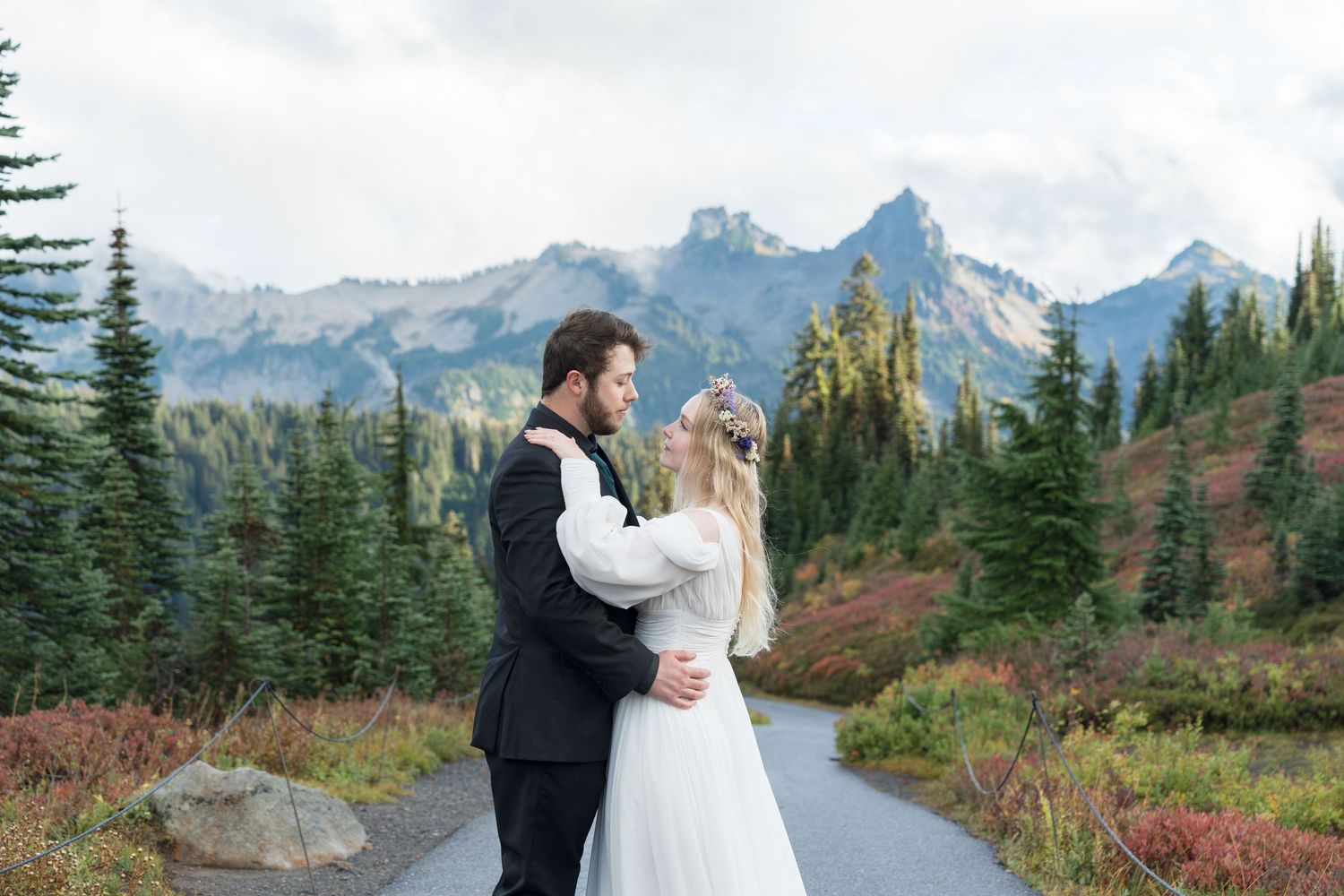 A couple in wedding attire stands atop a rocky outcrop with majestic mountain peaks and evergreen forest in the background.
