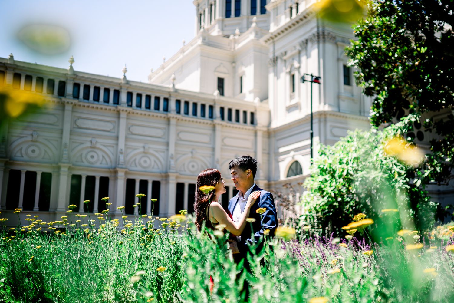 Couple embraces in front of a white cathedral building surrounded by wildflowers and green foliage taken by Melbourne couples photographer.