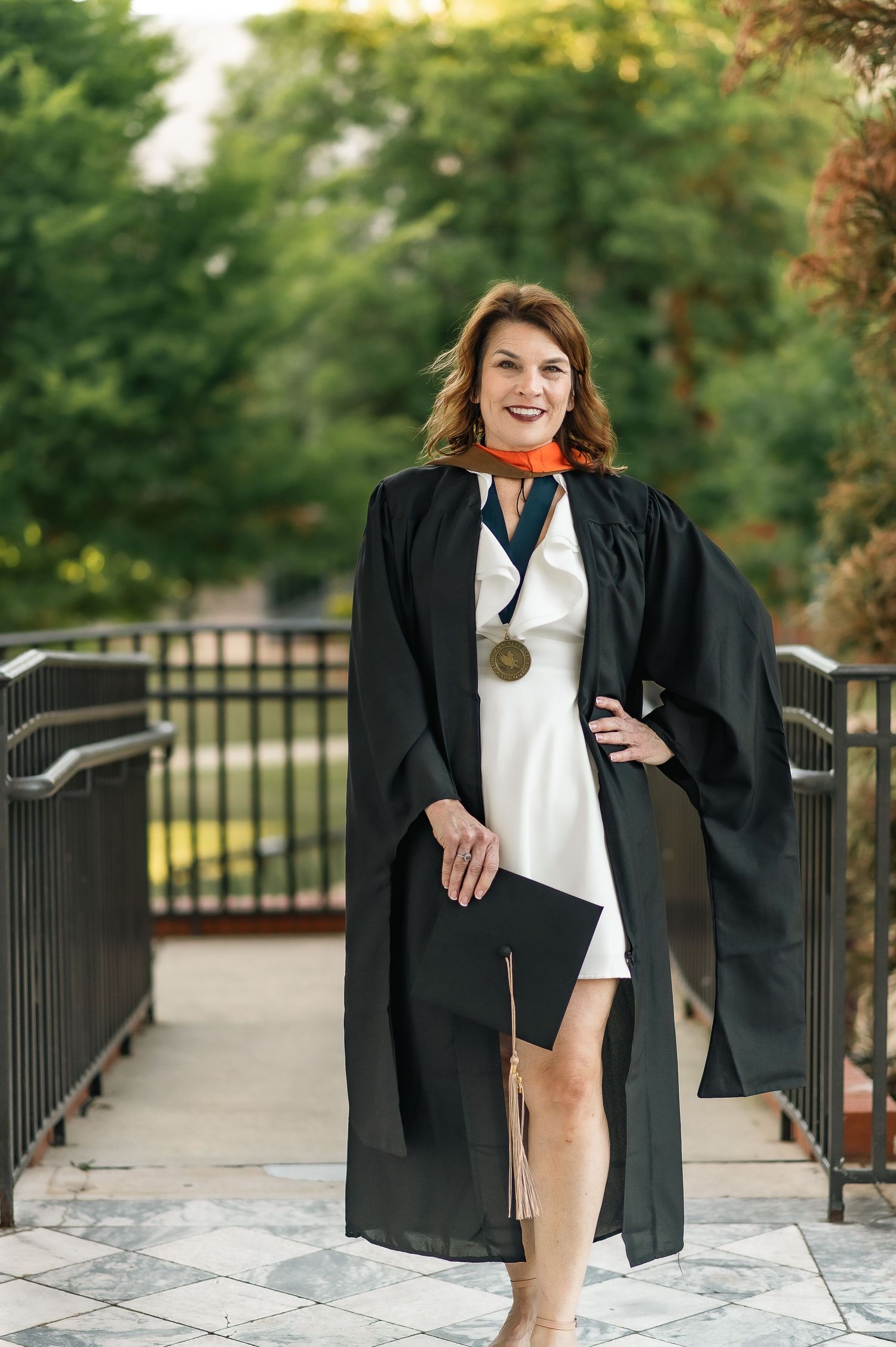Graduate wearing black cap and gown with white dress and medal poses on outdoor walkway in front of greenery.