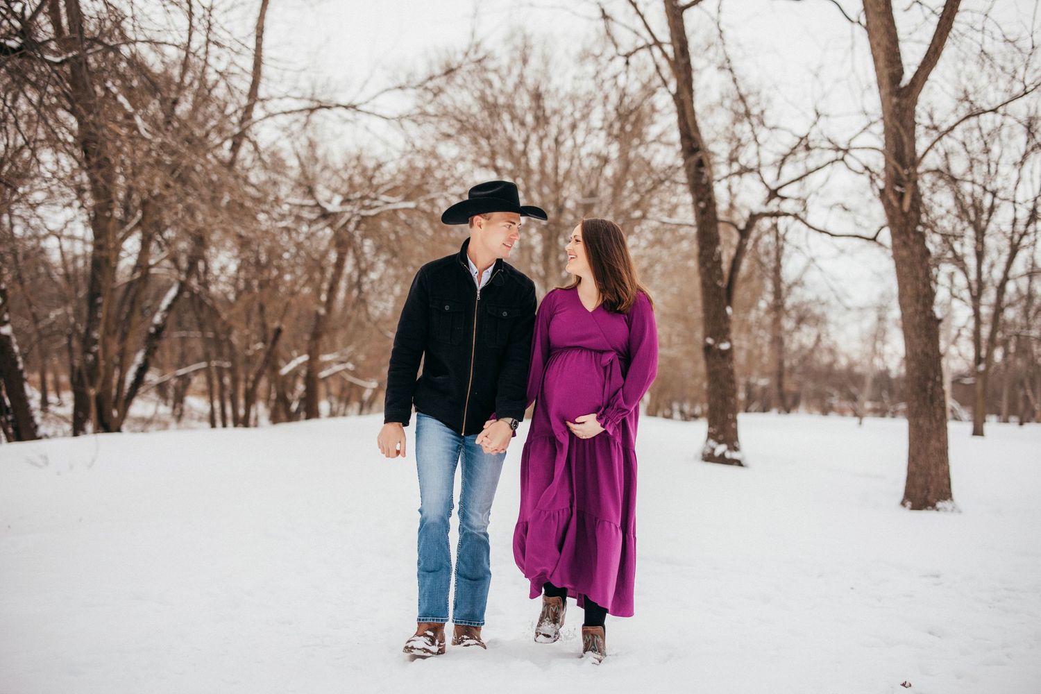 Couple walking through snowy winter scene with bare trees.