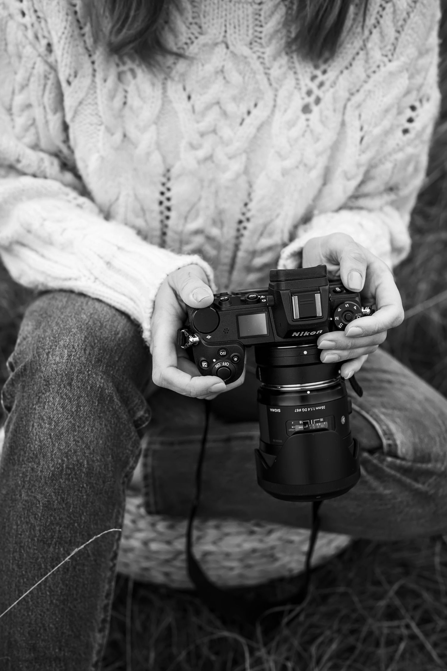 Black and white photo of hands holding a professional camera while wearing a cable knit sweater and jeans.
