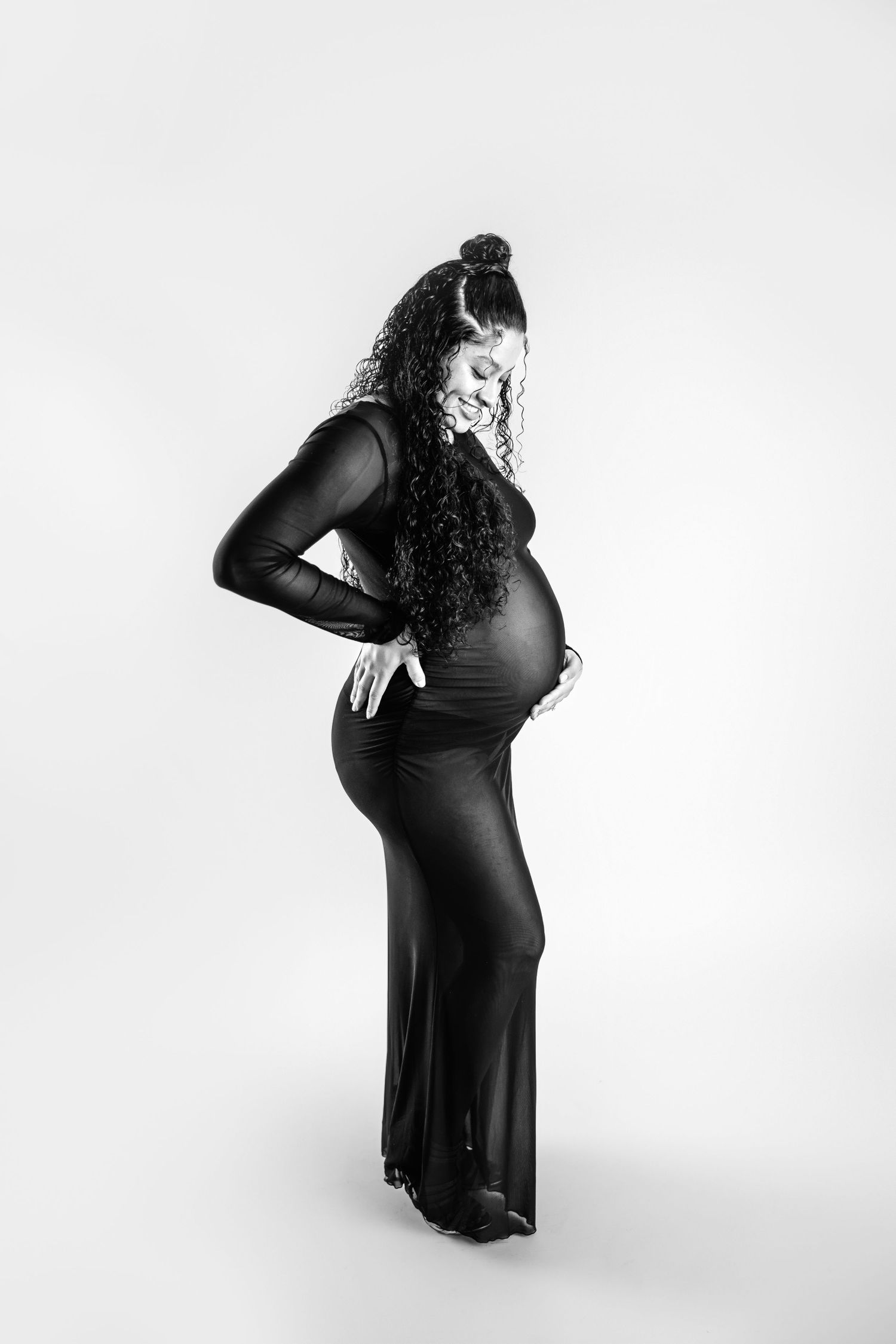 A silhouette of expectant mother in a long black dress poses elegantly against a white background in black + white. Maternity Photographer