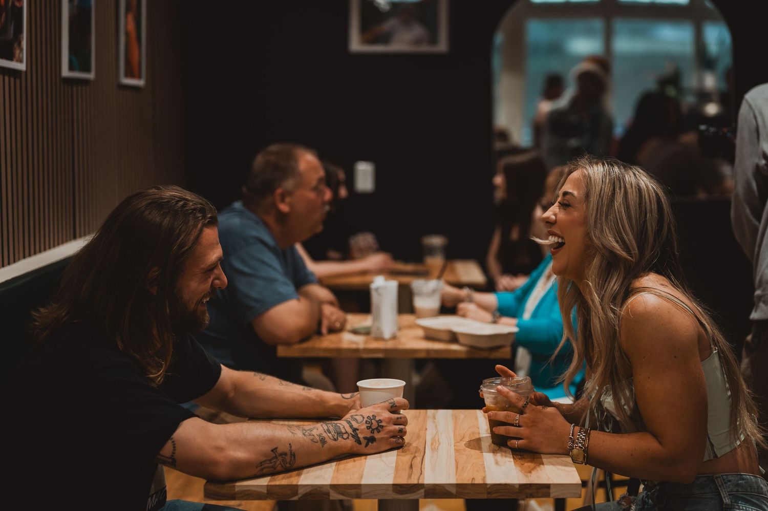 Two people share drinks and conversation at a wooden table in a dimly lit restaurant setting.