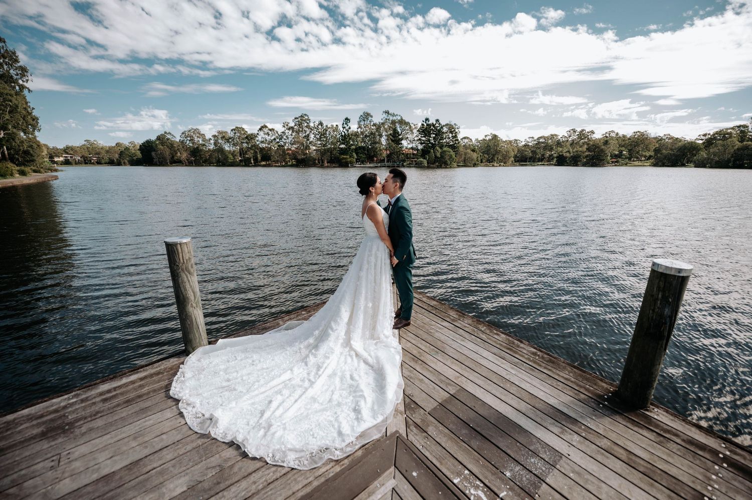 A couple stands together on a wooden dock overlooking a serene lake with trees and clouds in the background.