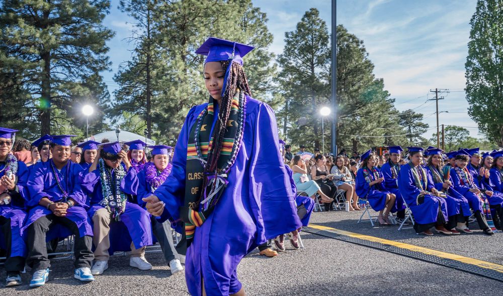 A graduation ceremony with students in bright blue caps and gowns seated outdoors under pine trees on a sunny day.