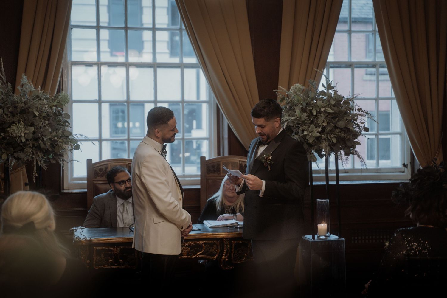 Two men in formal attire shake hands during a professional meeting in an elegant room with large windows and floral decor.