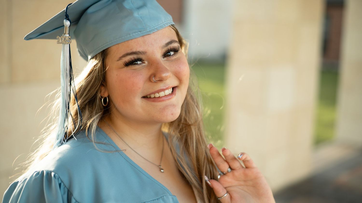 A graduate wearing light blue cap and gown smiles outdoors in natural sunlight.