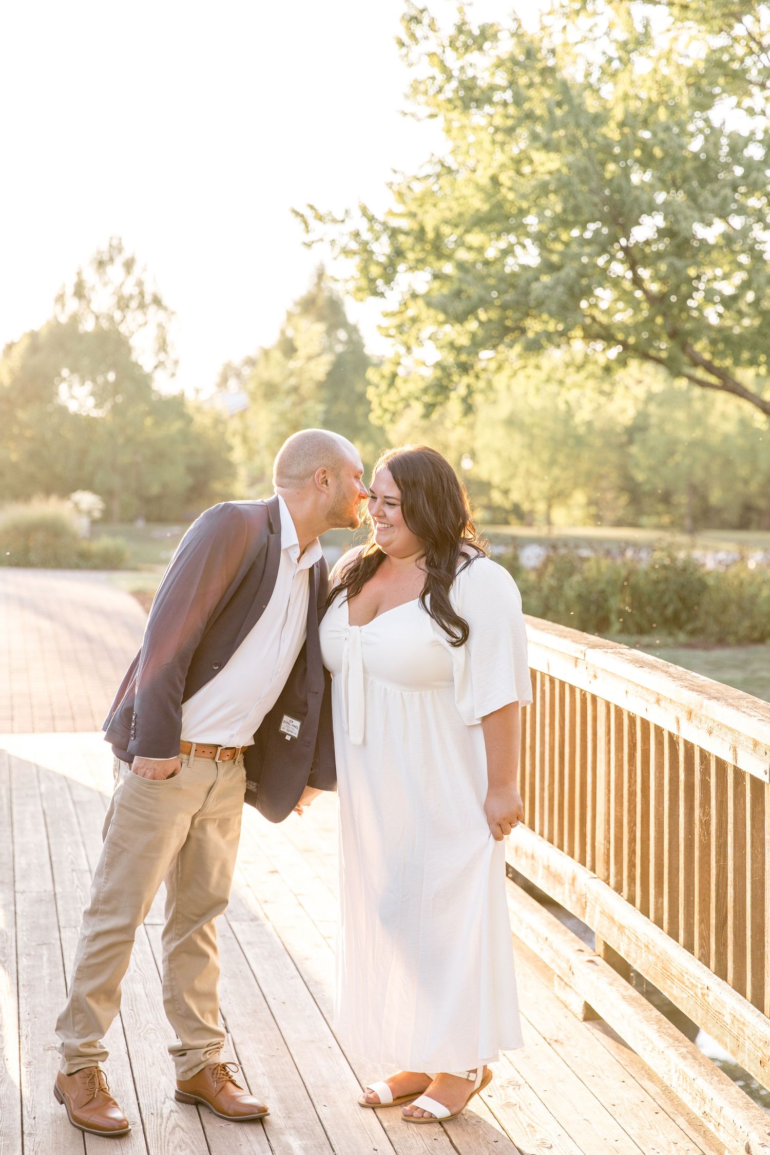 A romantic couple in white and tan stand close together on a wooden deck during golden hour sunset lighting.
