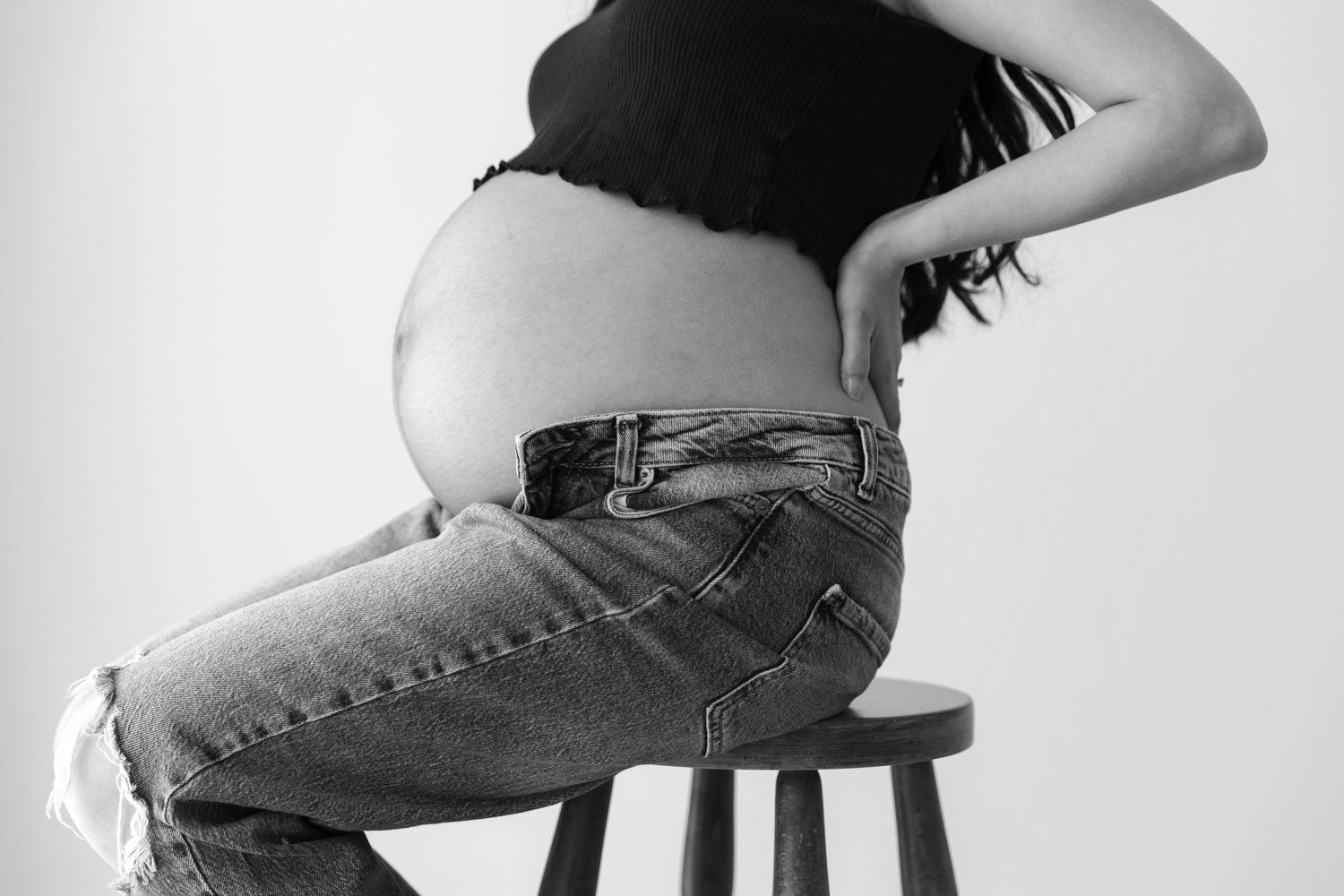 Black and white photo of a pregnant woman sitting sideways on a stool in torn jeans, showing her pregnant belly.