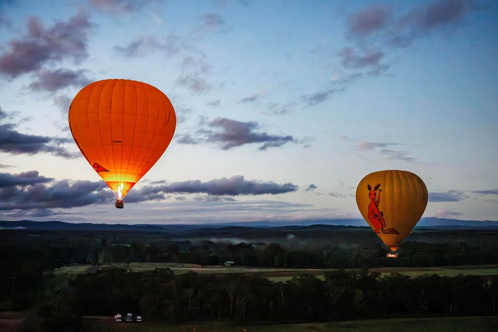 Two vibrant hot air balloons float over a lush green landscape at dusk under a cloudy sky. Cairns, Mareeba, Atherton, Australia