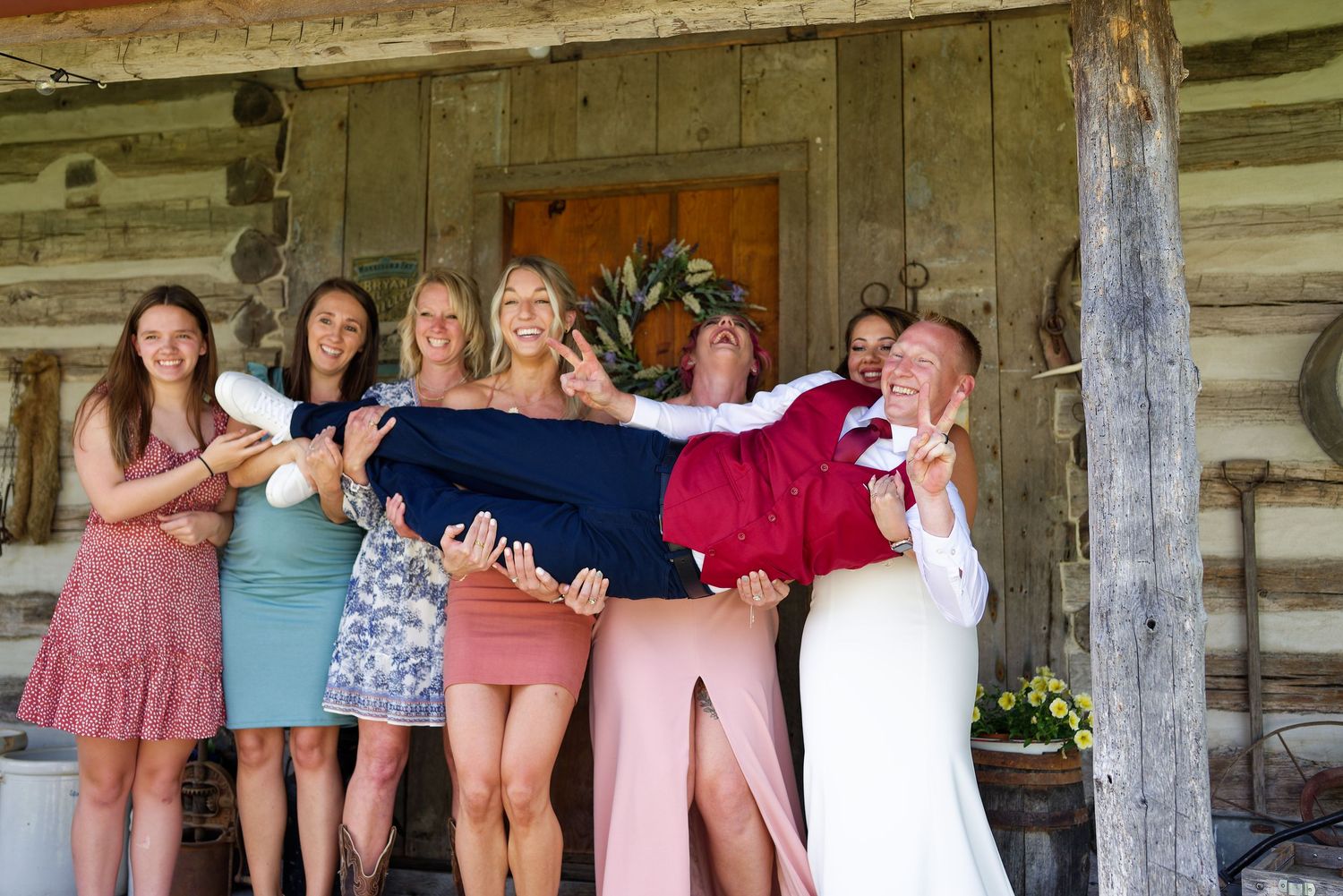 Wedding party lifts groom while bride stands beside rustic log cabin backdrop.