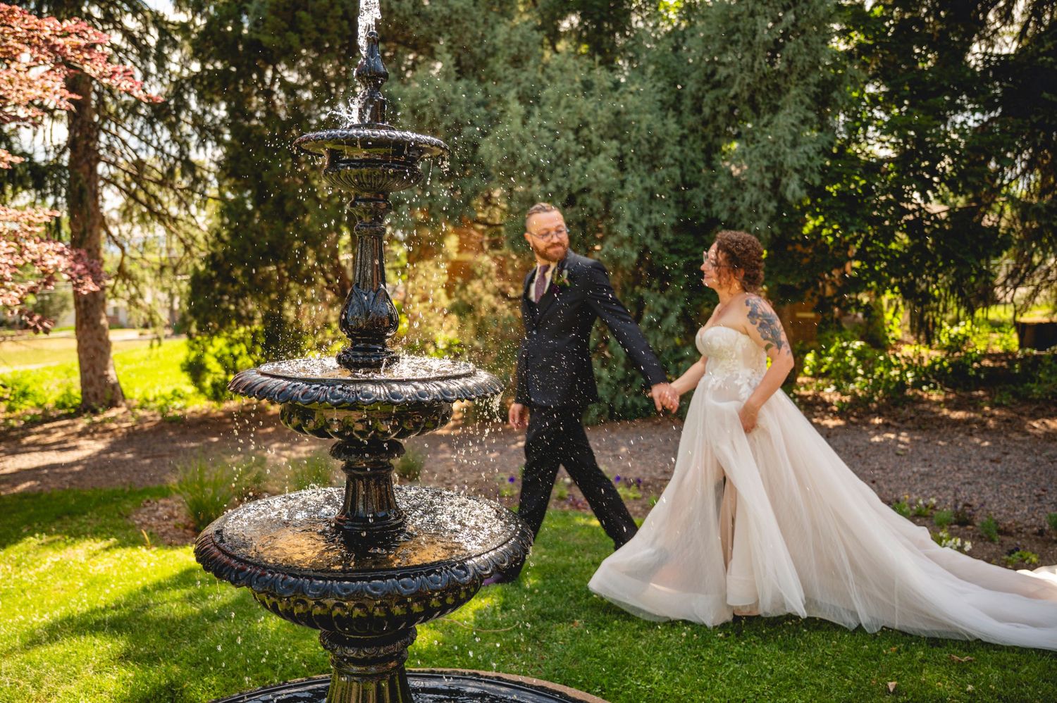 Bride in white gown and groom in suit walk hand in hand near ornate fountain in lush garden wedding setting.