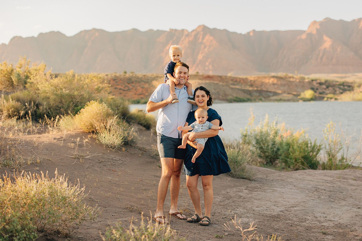 Family of four posing outdoors near a lake with mountain backdrop in warm golden hour light.