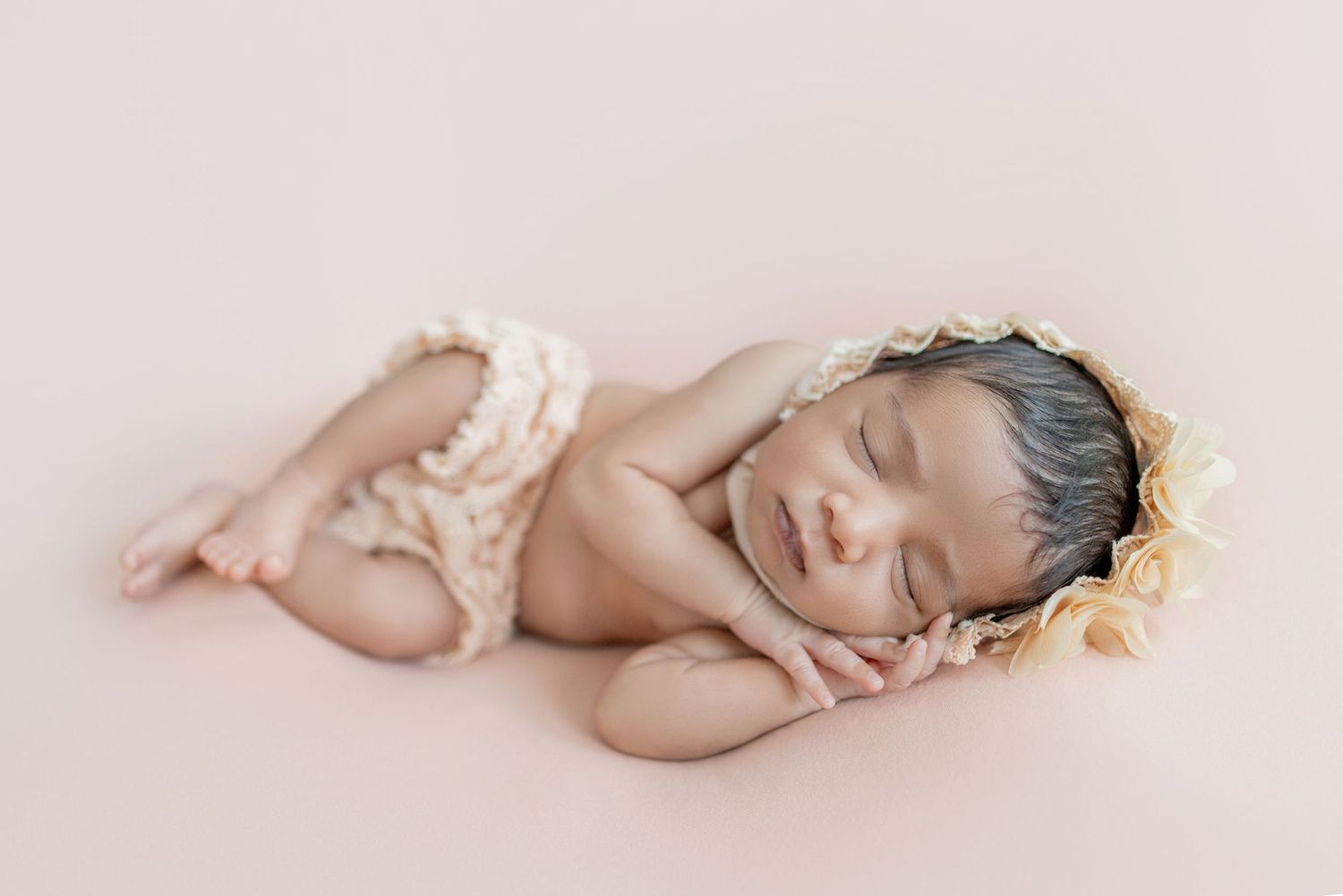 Sleeping newborn baby wearing a floral bonnet and ruffled bloomers, posed on a soft pink background.