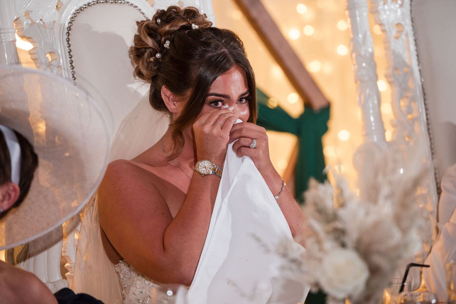 Emotional bride in strapless gown wiping tears with white cloth at wedding reception with fairy lights.