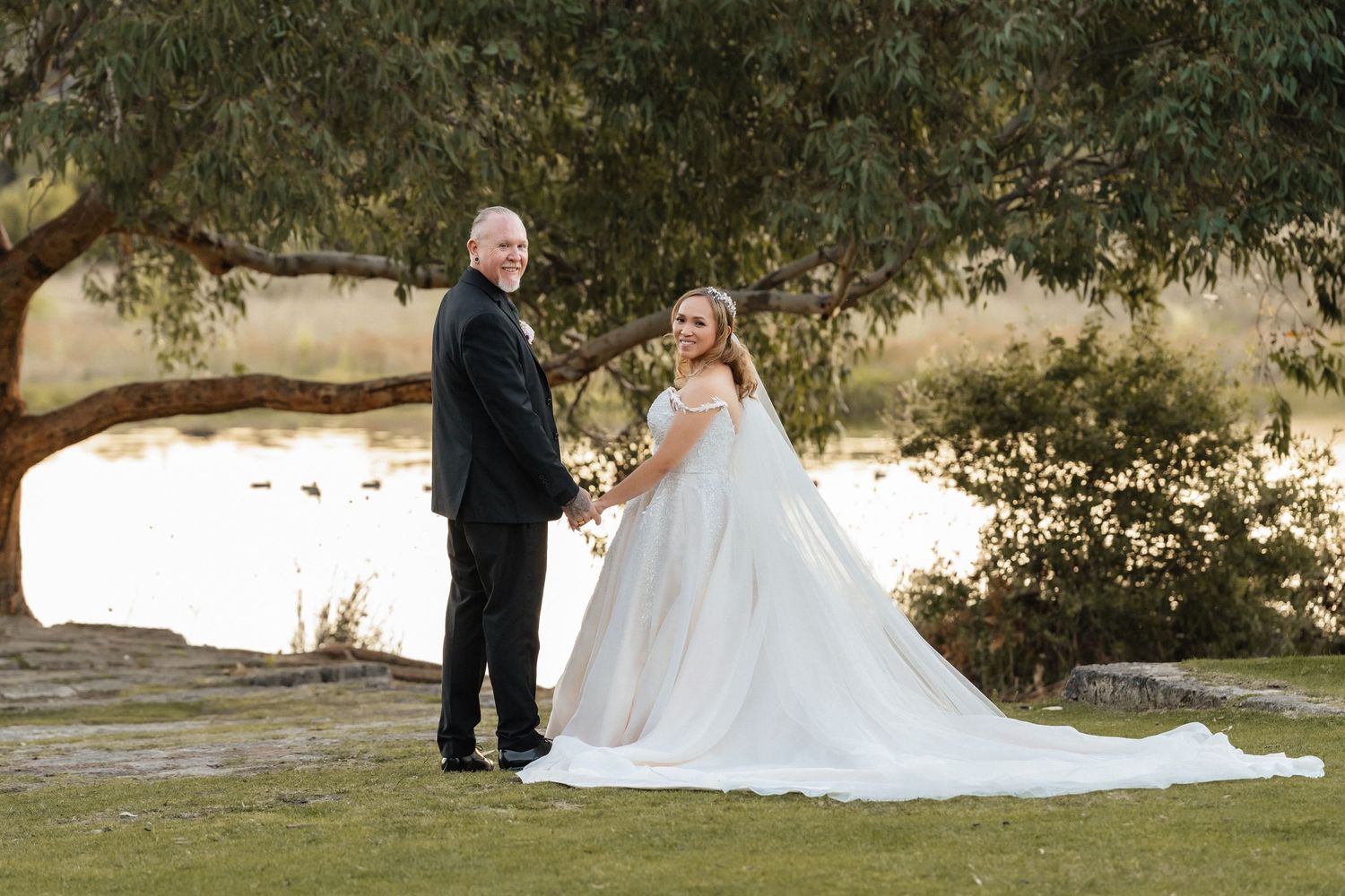 Bride and groom holding hands under a large tree by a pond, bride wearing a flowing white gown with a long train.