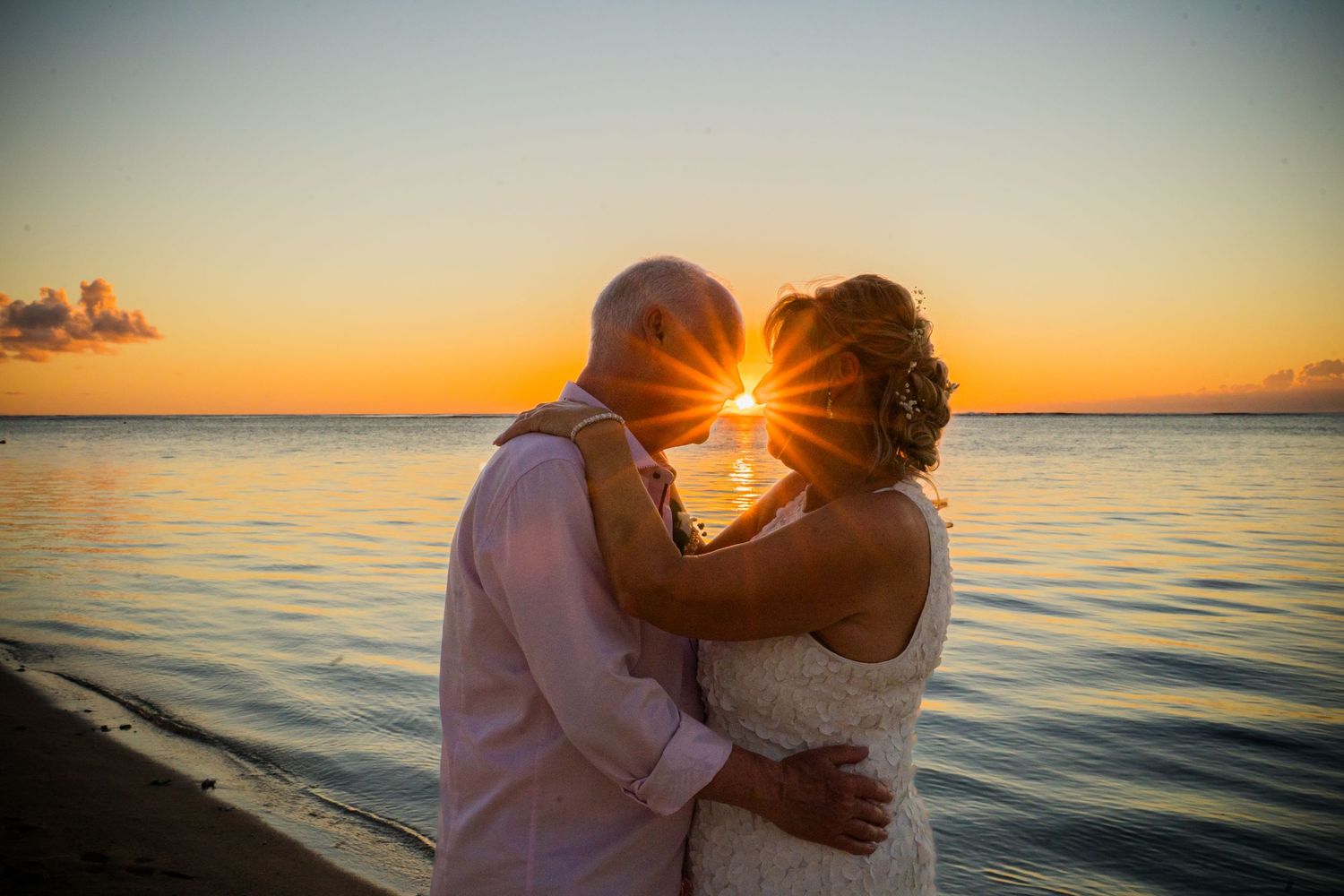 Couple embracing at sunset on a beach in wedding attire, with golden sun rays between them over calm ocean waters.