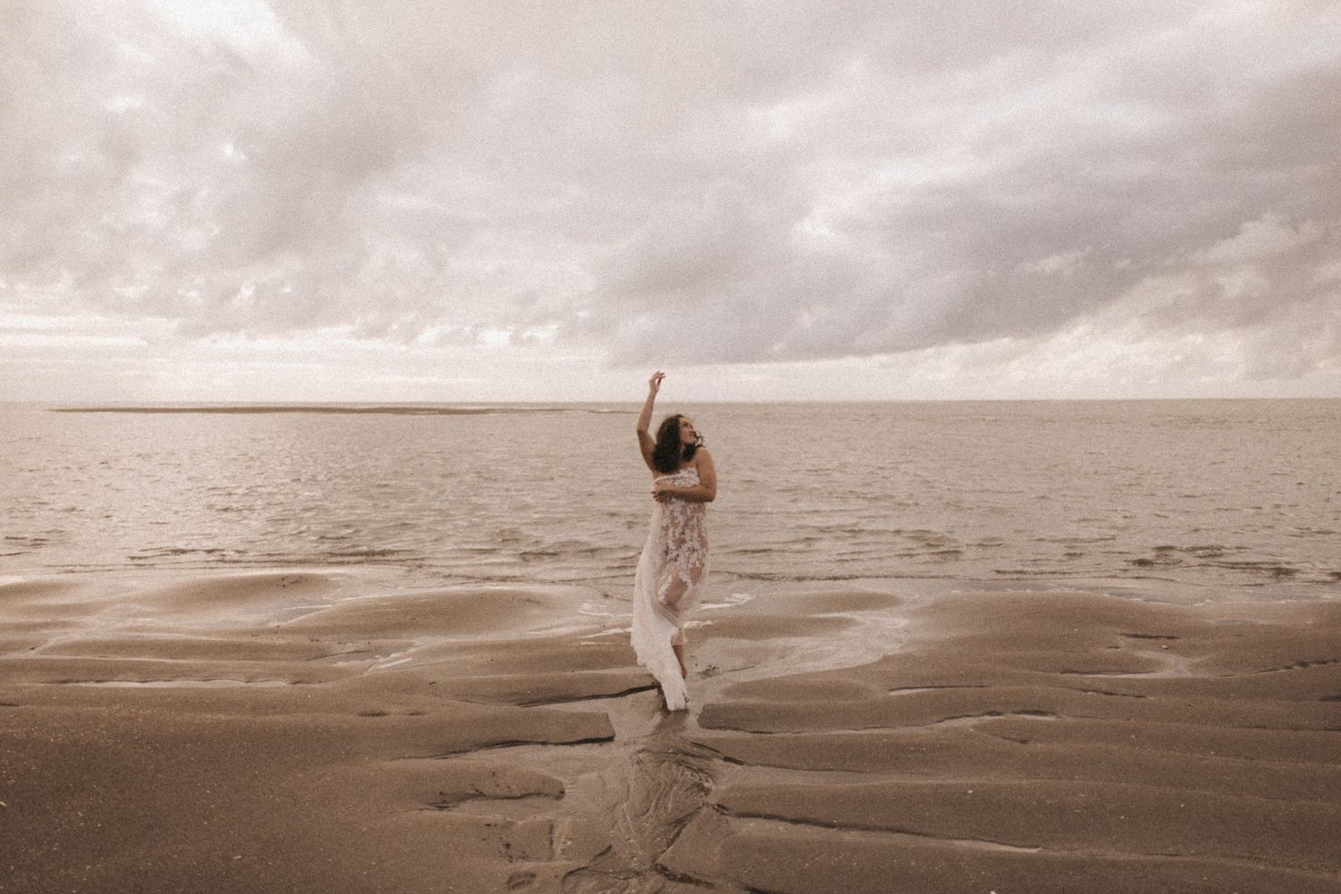 A woman in white stands on a sandy beach with arms raised, under a dramatic cloudy sky near the ocean.