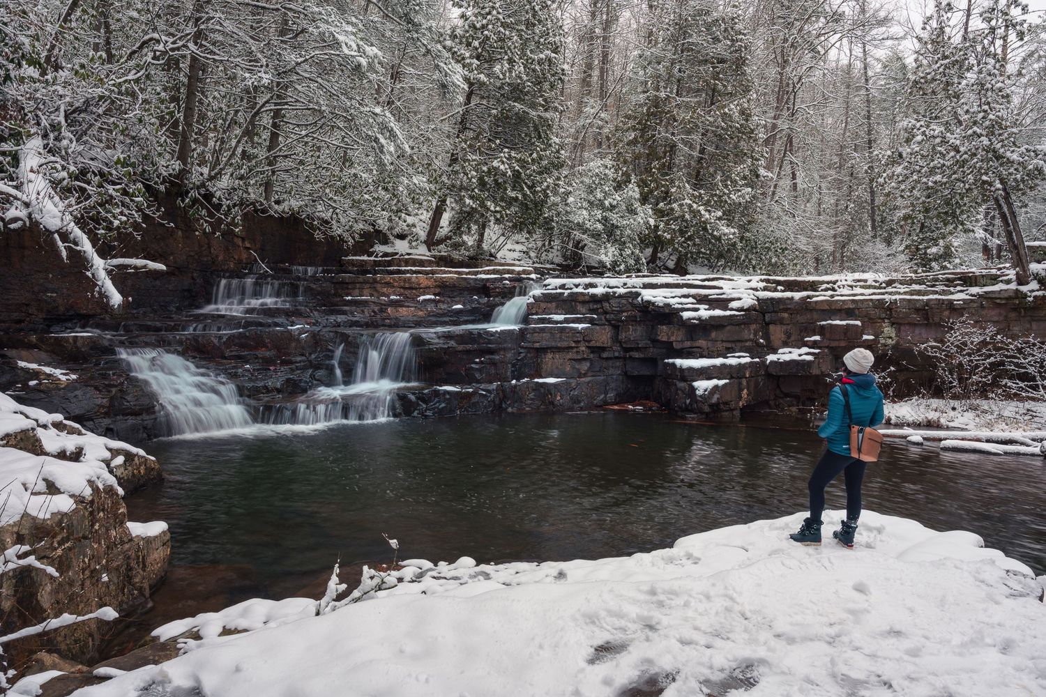 Hiker in winter gear stands beside a snow-covered waterfall cascading over rocky ledges in a forest.