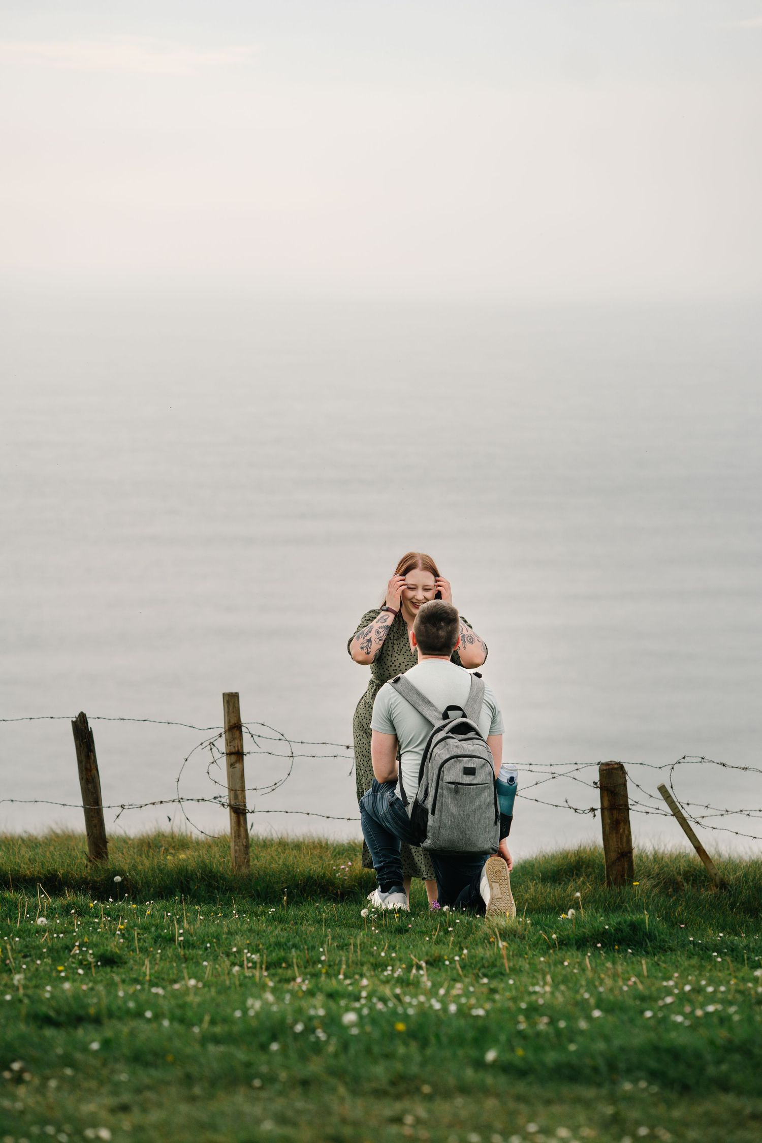 Couple sharing a romantic moment on a grassy coastal cliff, with barbed wire fence and ocean in the background.