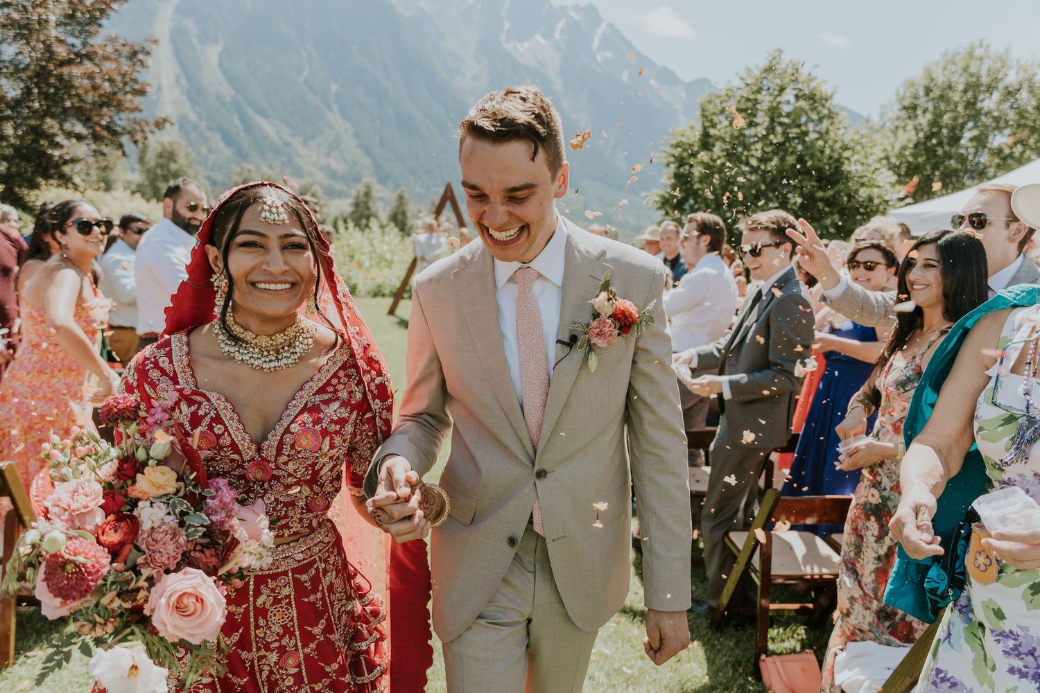 A couple in traditional South Asian wedding attire poses together in a sunny outdoor garden setting.