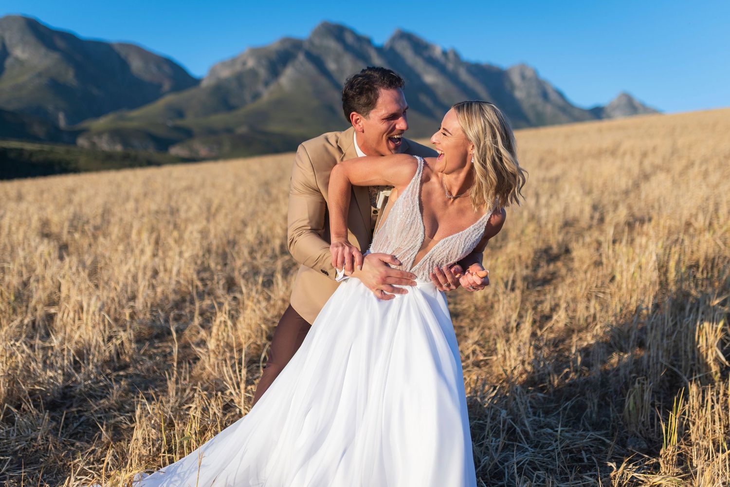 Couple embracing in golden wheat field with mountain backdrop on their wedding day.