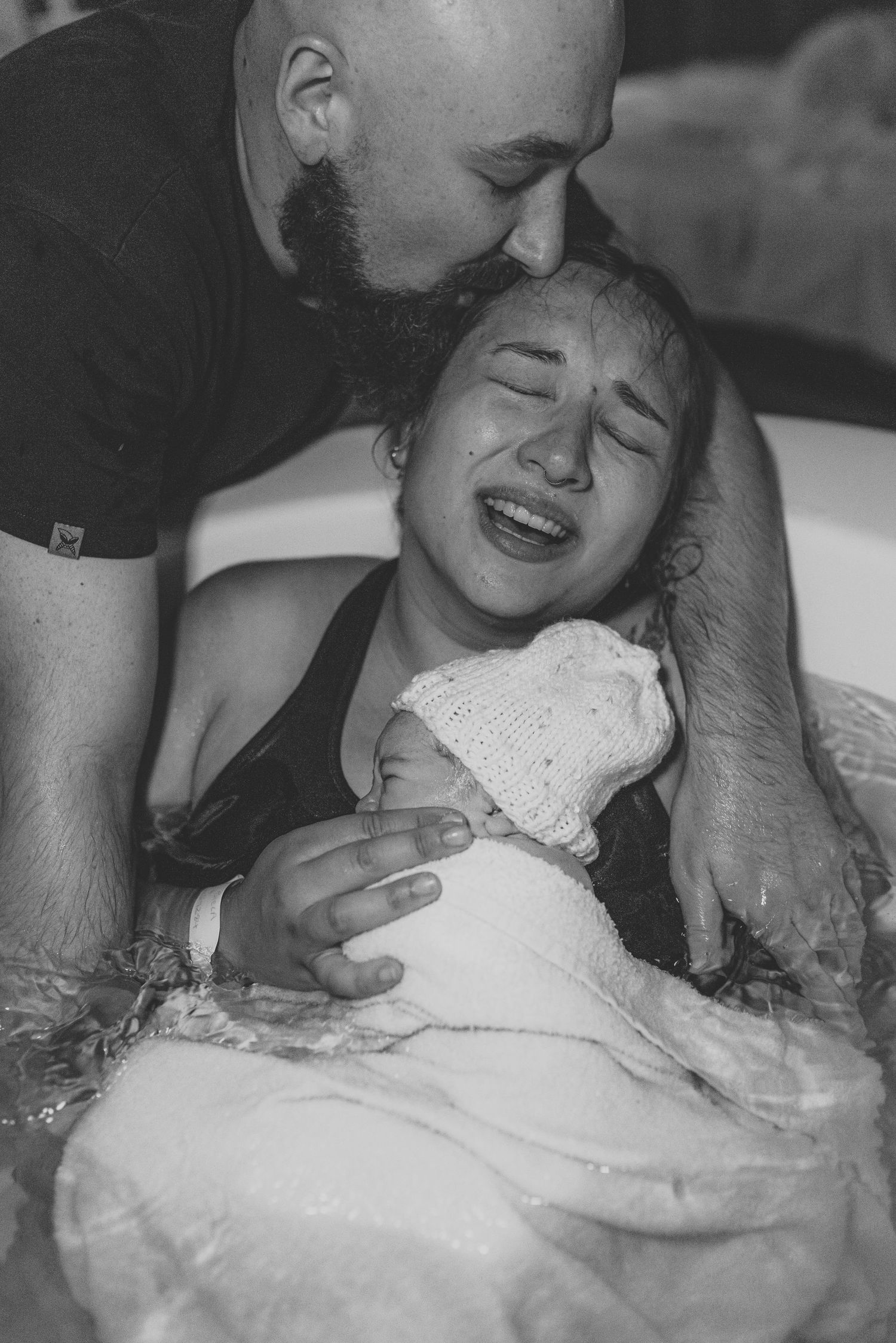 Black and white photo of a woman crying tears of joy holding her newborn baby in a birthing tub, partner kissing her head.