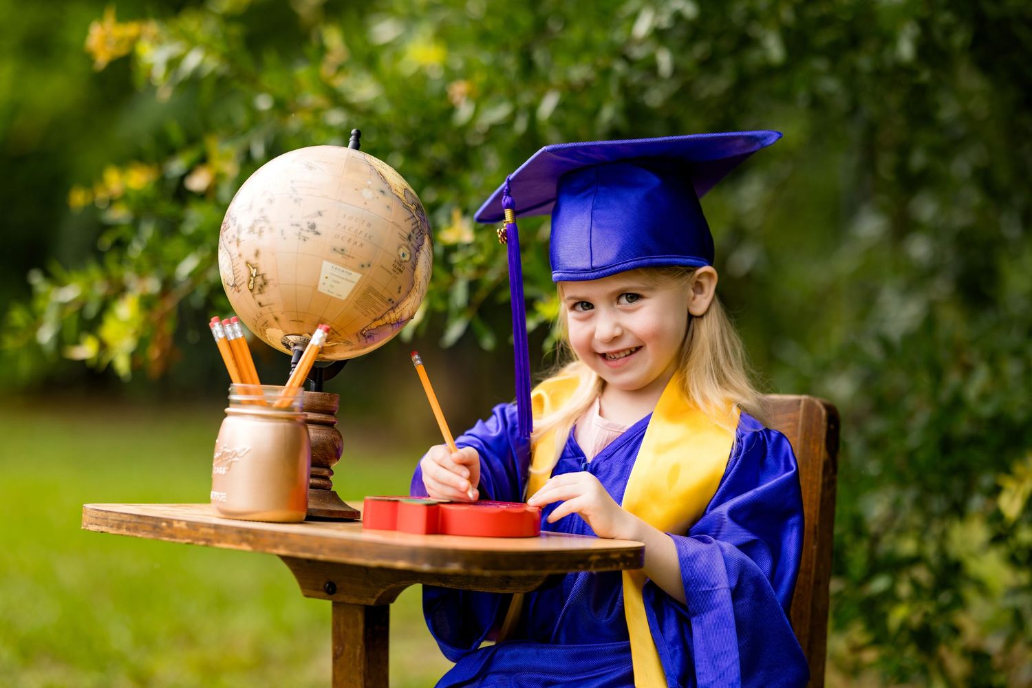 Young girl in blue graduation cap and gown smiles at desk with globe and pencils outdoors.