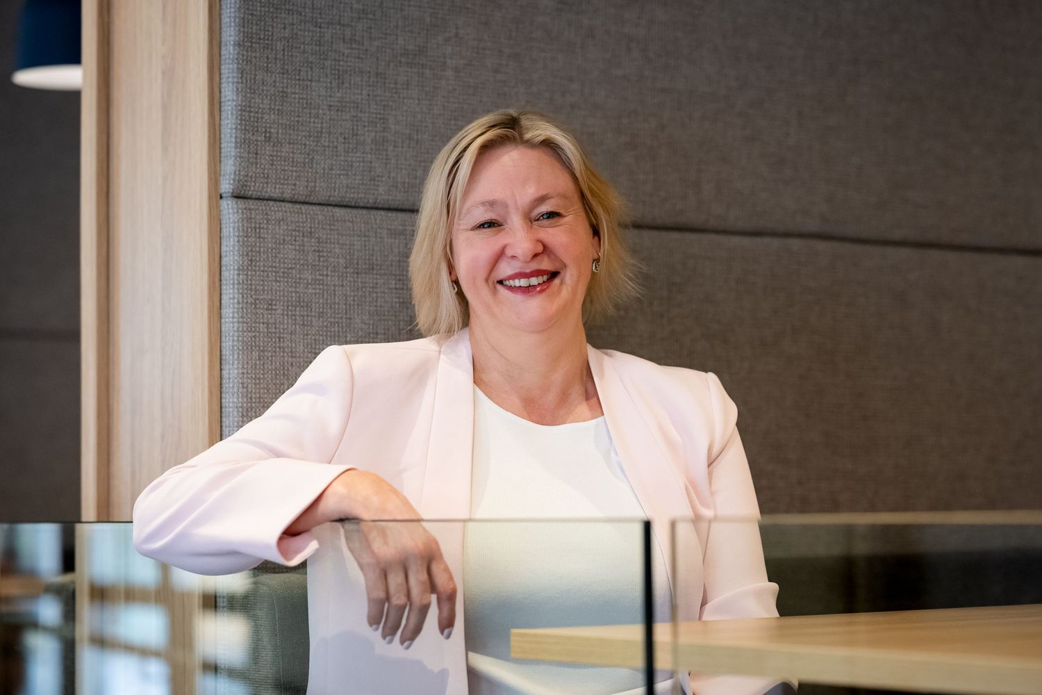 Smiling professional woman in white blazer leaning on glass desk in modern office setting.