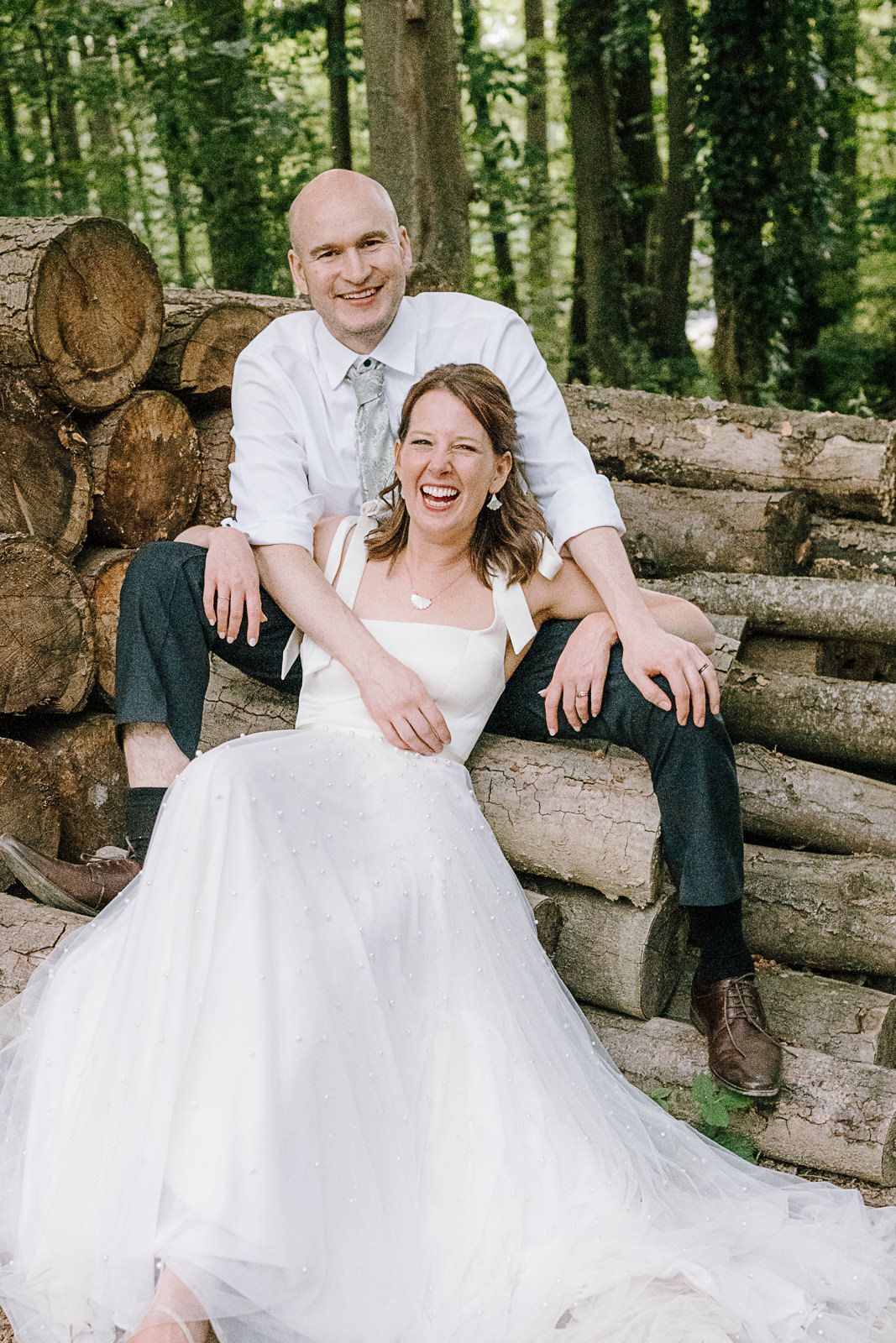 Happy bride and groom sitting on stacked logs in a lush forest, smiling joyfully in wedding attire.